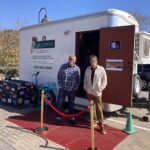 Two men stand in front of the "Mobile Studio USA" trailer, which is set up with a red carpet and small crowd control posts, giving the appearance of an event setup. The trailer door is open, showing part of the interior recording setup, suggesting a promotional or open house display.