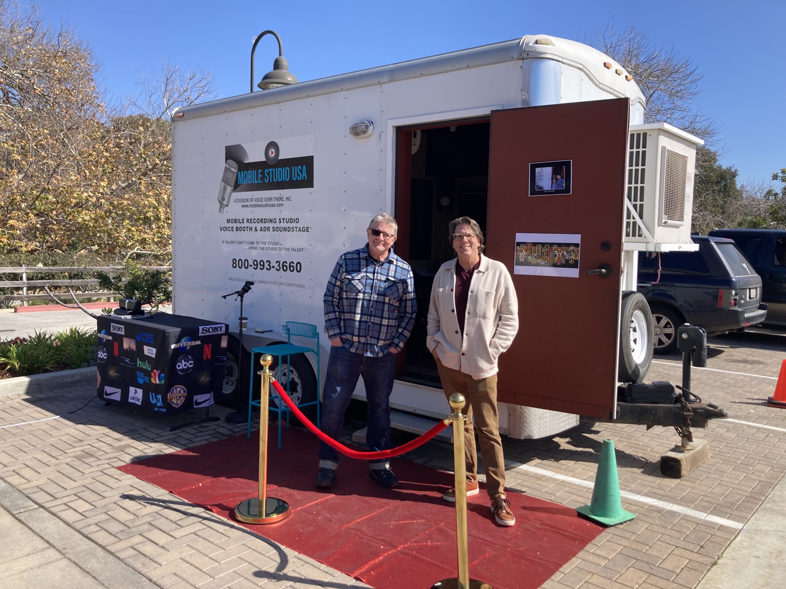 Two men stand in front of the "Mobile Studio USA" trailer, which is set up with a red carpet and small crowd control posts, giving the appearance of an event setup. The trailer door is open, showing part of the interior recording setup, suggesting a promotional or open house display.